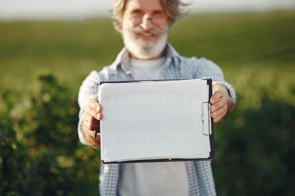 Bearded senior man with glasses holding a clipboard in a green field on a sunny day.