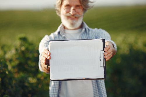 Free Bearded senior man with glasses holding a clipboard in a green field on a sunny day. Stock Photo