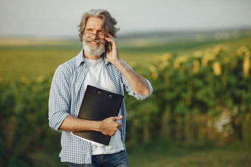 Free Elderly man with beard and glasses talking on phone outdoors in sunflower field. Stock Photo