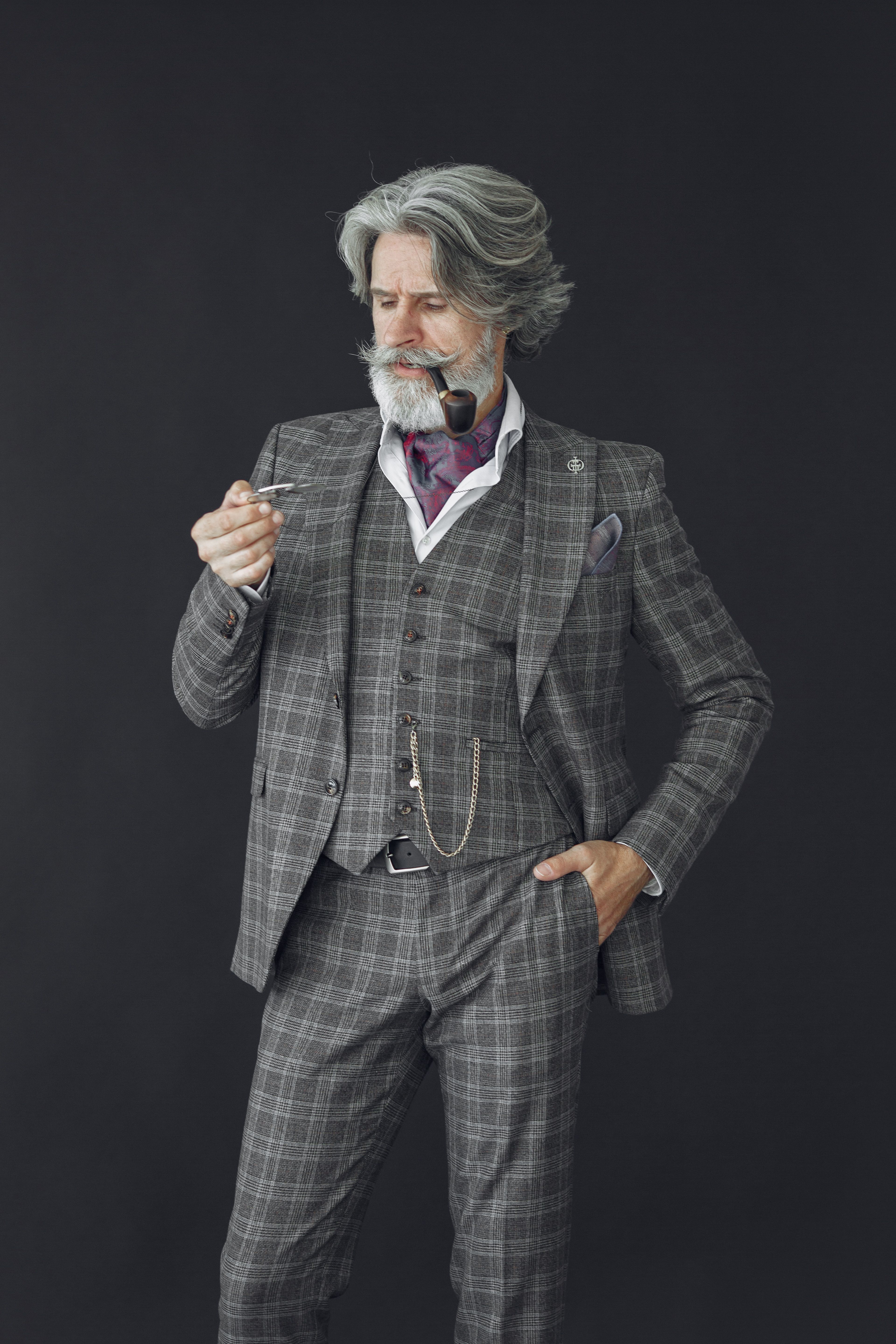 A refined senior man with gray hair poses confidently in a checkered suit holding a tobacco pipe.