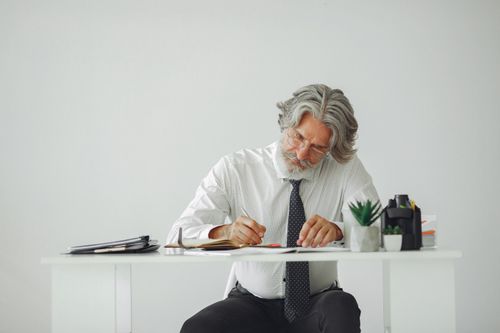 Free Elderly businessman with gray hair writes at a desk in a modern office. Stock Photo