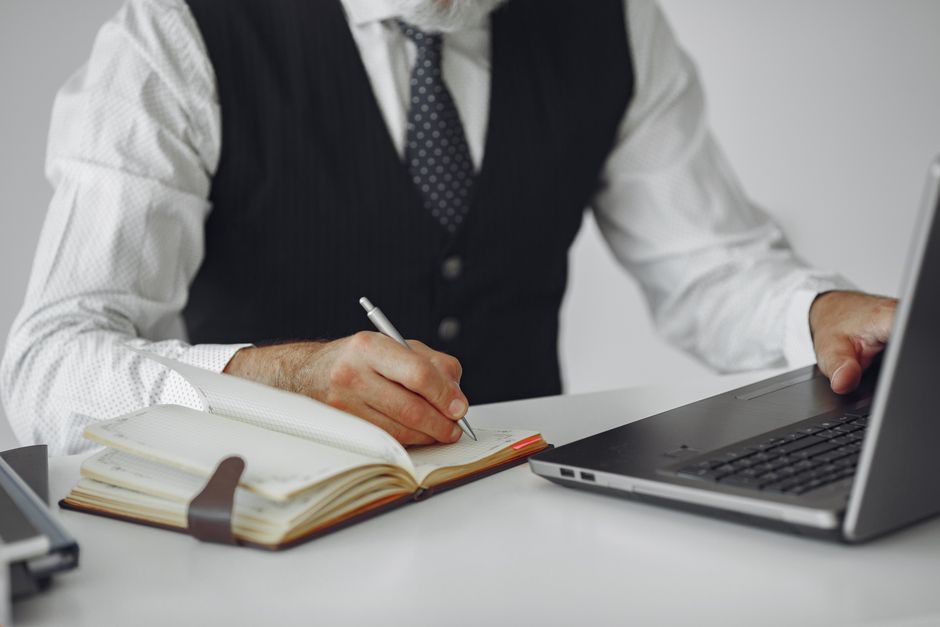 Business professional writing notes while working on a laptop at an office desk.