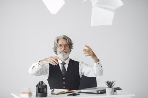 Free Elderly man in formal attire throwing papers at modern desk, symbolizing stress or creativity. Stock Photo