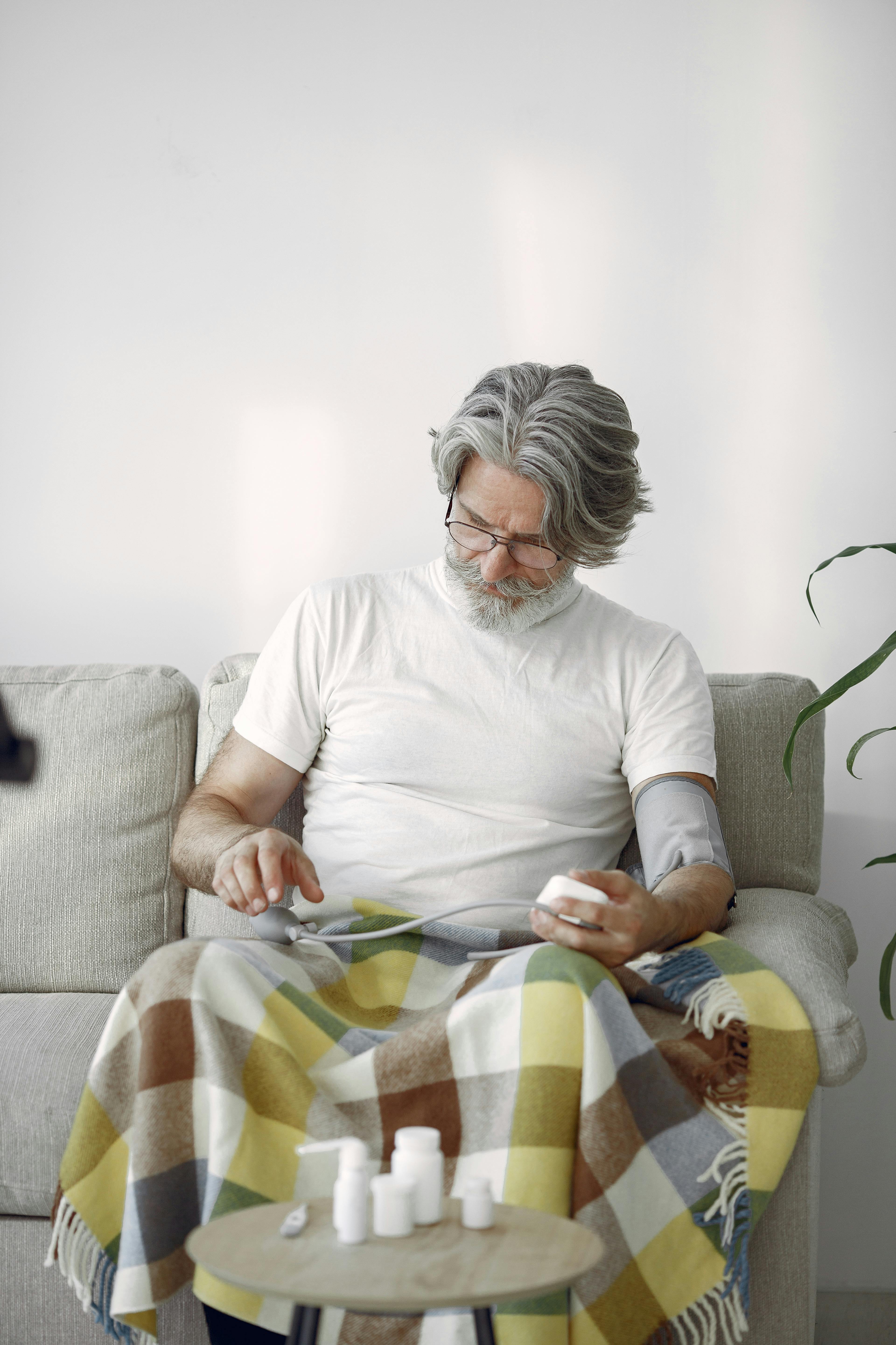 Elderly man sitting on sofa with blanket, monitoring blood pressure with medication bottles.