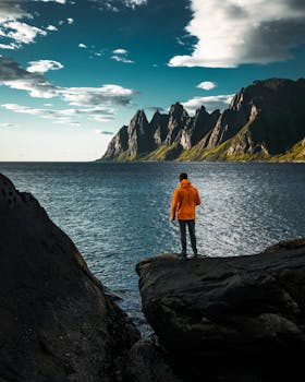 A person in an orange jacket stands on rocks overlooking a stunning fjord in Norway.