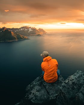 Man in orange jacket sits on cliff, overlooking a serene ocean and mountain landscape at sunset.