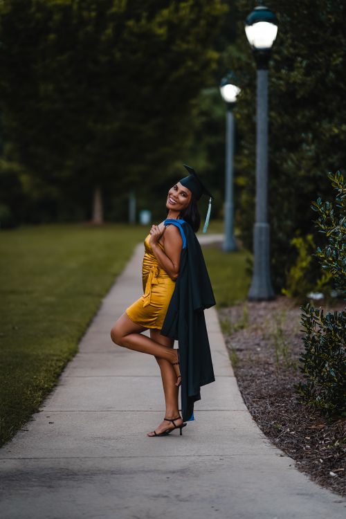 Free Full body of smiling female wearing elegant dress and graduation cap looking at camera while standing on narrow walkway with raised leg and cape on shoulder Stock Photo