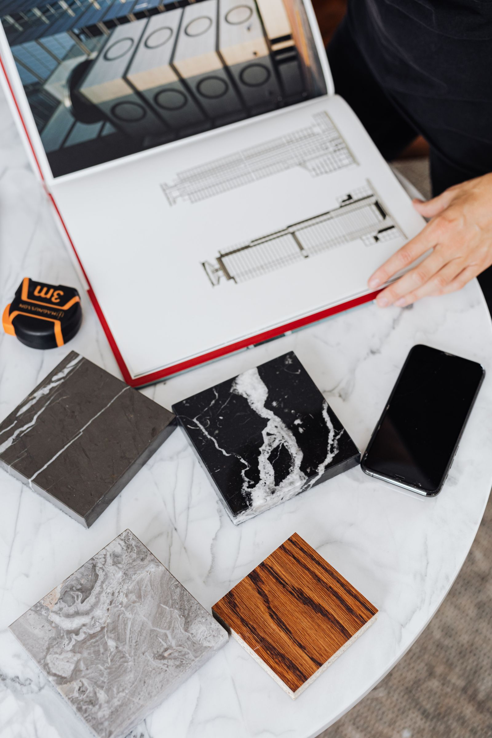 Overhead view of architectural plans, stone and tile samples, a tape measure, smartphone, and a person reviewing materials.