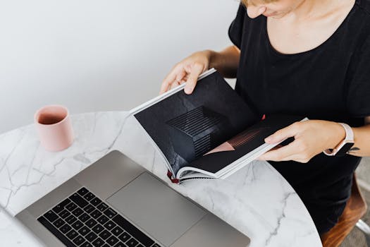 A woman reads a book at a marble table with a laptop and coffee cup in a modern workspace.