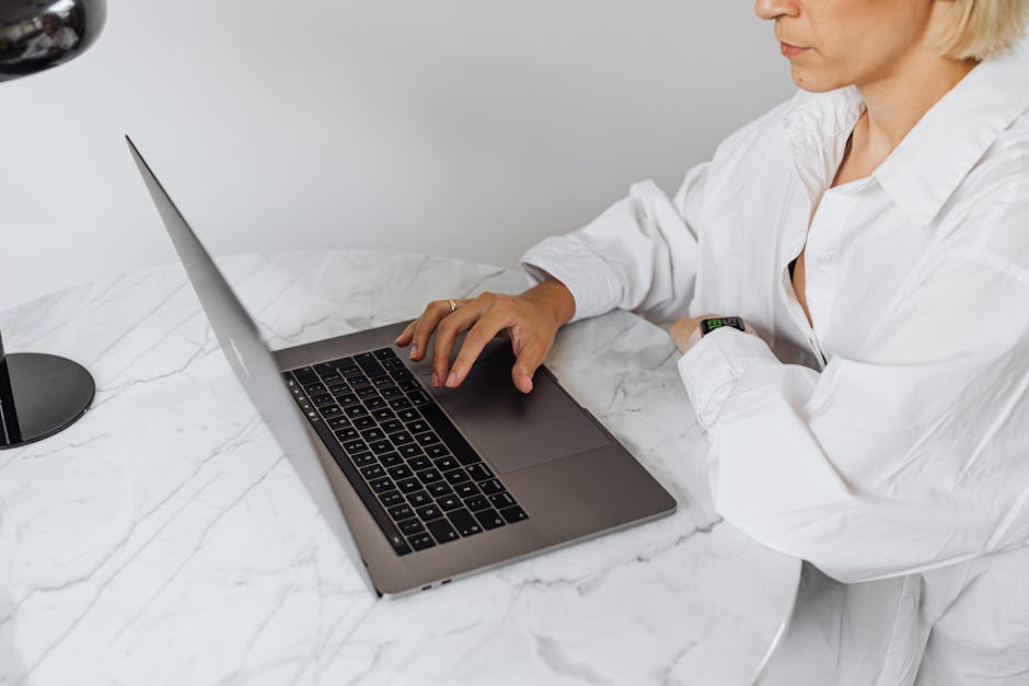 Caucasian woman in a white shirt working on a laptop at a marble table indoors. - My Strategy for Crafting a Resume for Remote Jobs.