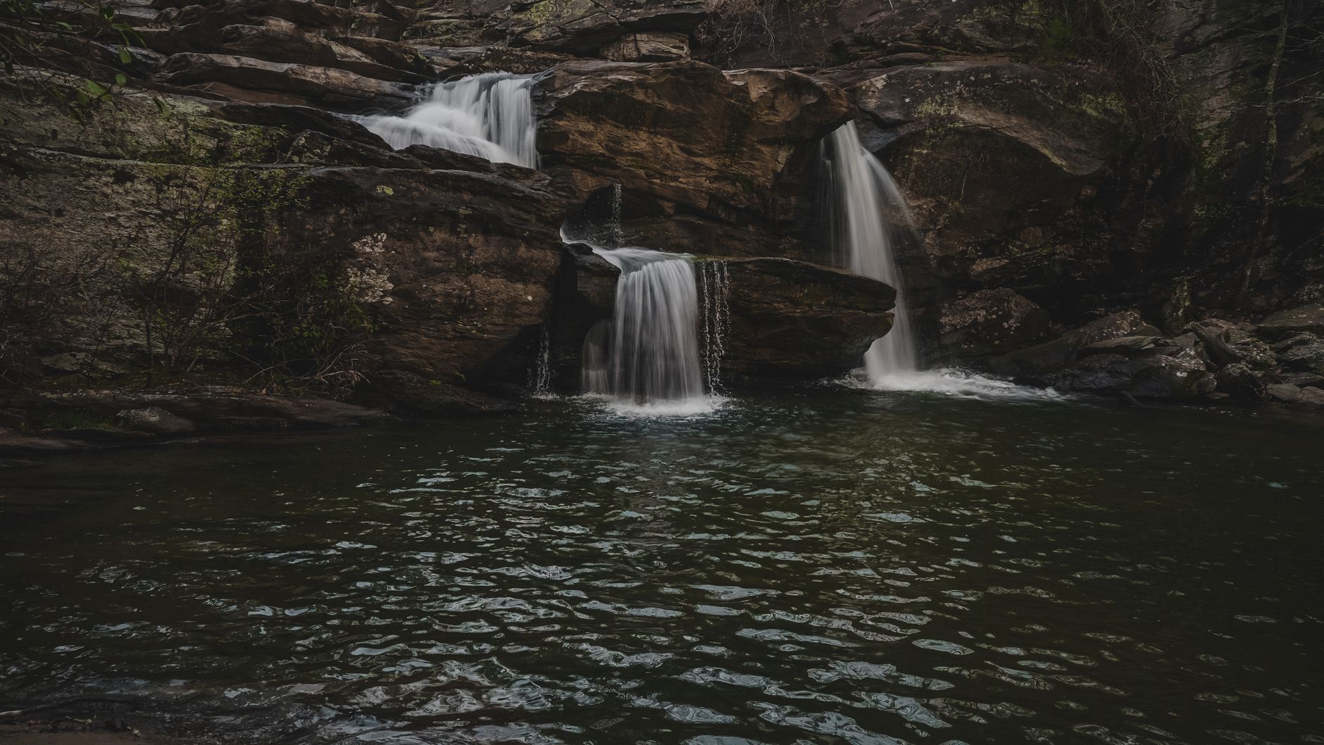 A vivid waterfall drops over layered rock into a forested pool, with bright daylight and dense green plants surrounding the scene.