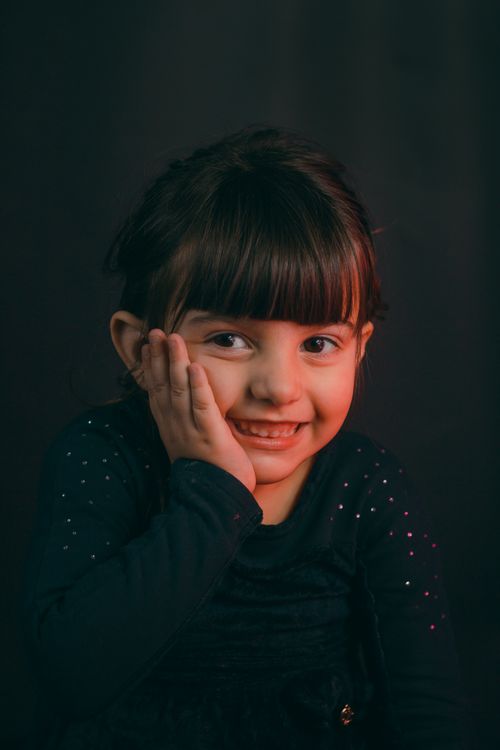 Free Adorable happy little girl with dark hair in casual clothes touching cheek and smiling against black background Stock Photo