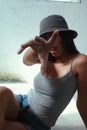 Sensual young female in jeans shorts and top showing tongue and reaching out hand towards camera while sitting on floor in sunlight