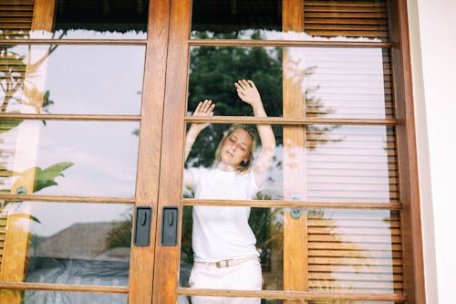 Woman in White Shirt Standing Beside Brown Wooden Framed Glass Windows