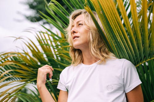 Woman in White Crew Neck T-shirt Standing Near Palm Leaves