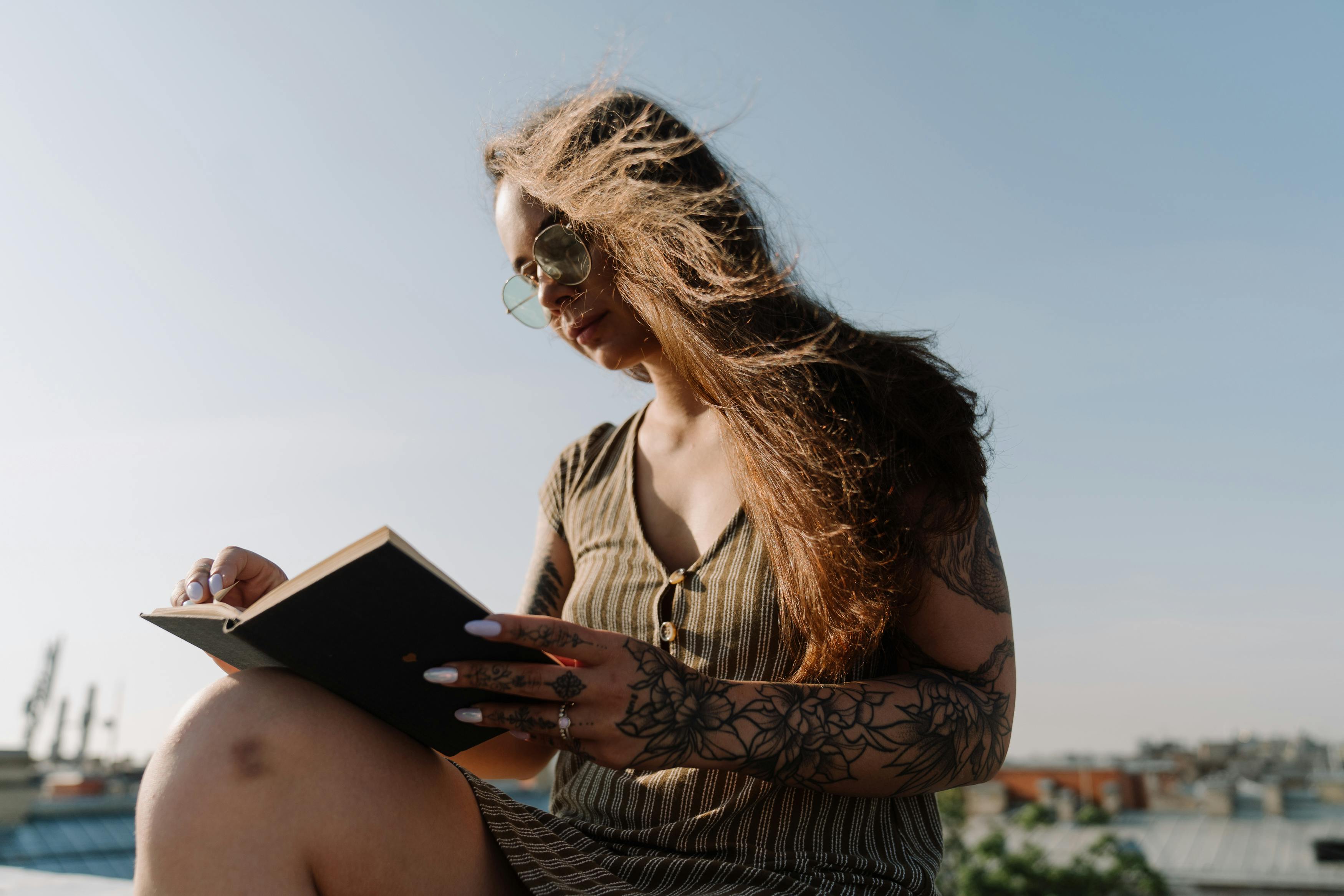 A tattooed woman enjoying a sunny day reading on a rooftop.