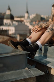 Tattooed legs with black boots resting on a rooftop in an urban cityscape.