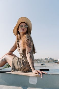 Stylish woman with tattoos enjoys a sunny day on a rooftop, embodying urban summer vibes.