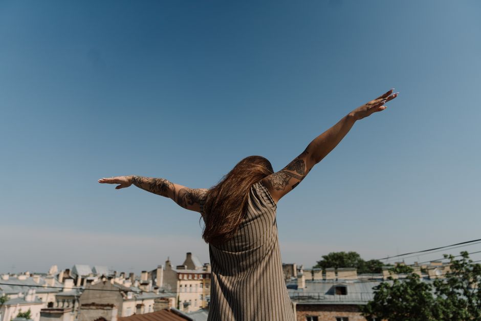 A woman with tattoos stands freely on a rooftop, arms open against a clear blue sky and cityscape background