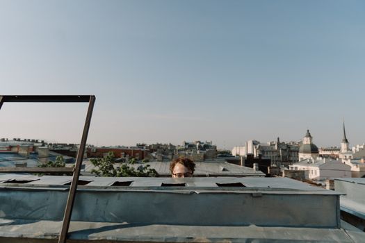 A man peeking over a rooftop with a cityscape in the background under a clear sky.