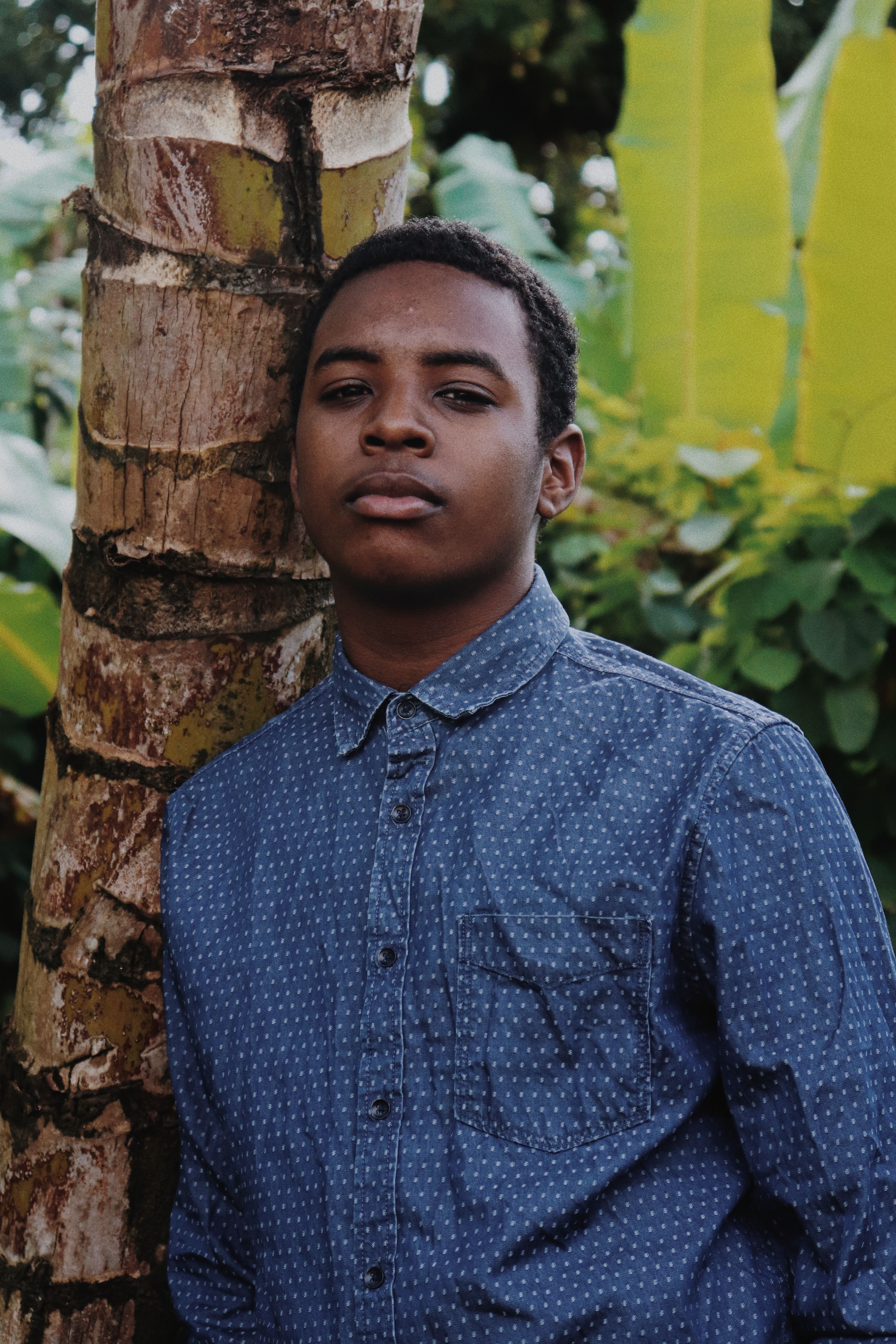 Portrait of a young man in a denim shirt leaning against a tree, surrounded by lush greenery.