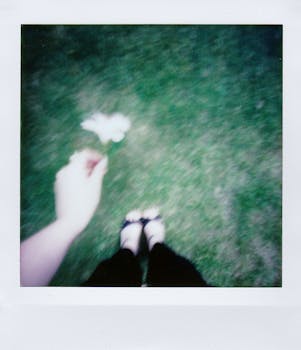 A defocused Polaroid photo capturing a hand with a flower, feet visible on grass.