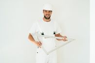 Happy carpenter in white outfit holding a large square ruler indoors.