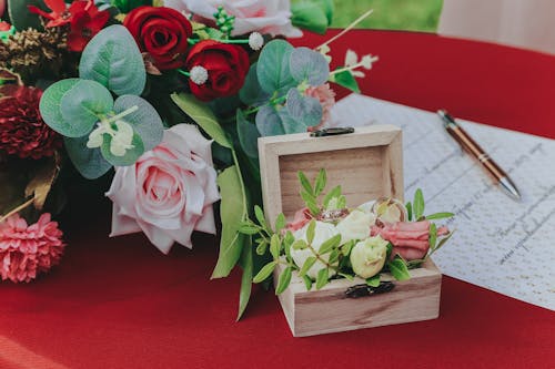 From above of small box with wedding rings near bouquet of flowers and paper on red table