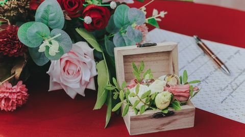 From above of small box with wedding rings near bouquet of flowers and paper on red table