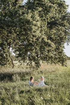 两个年轻女孩坐在一棵大树下的草地上，享受着户外阳光明媚的一天