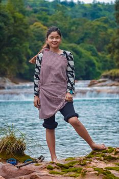 A young woman stands confidently by a flowing river in a lush forest setting.