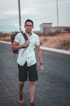 Casual portrait of a young man walking on a deserted road with a backpack and eyeglasses.