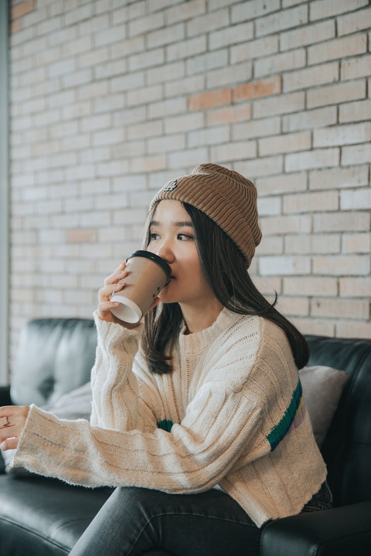 Dreamy Asian Woman In Stylish Wear Drinking Coffee On Sofa