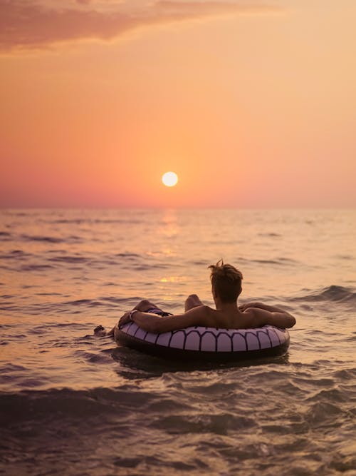 Back view of unrecognizable male tourist lying in inflatable ring on wavy sea while contemplating bright sky with sun in evening