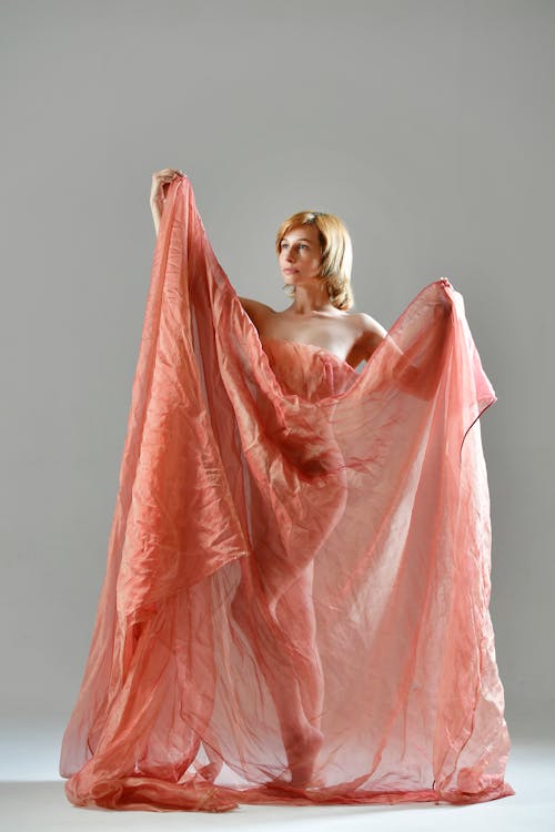 Young female covering nude body with pink organza and looking away on grey background
