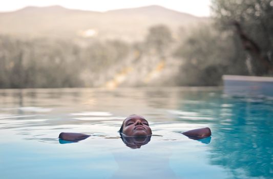 A person floating calmly in a serene pool with olive trees and hills in the background.