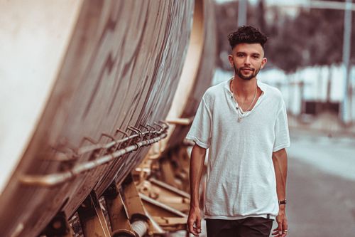 Free A man in a casual white t-shirt walking beside industrial structures, showcasing modern street fashion. Stock Photo