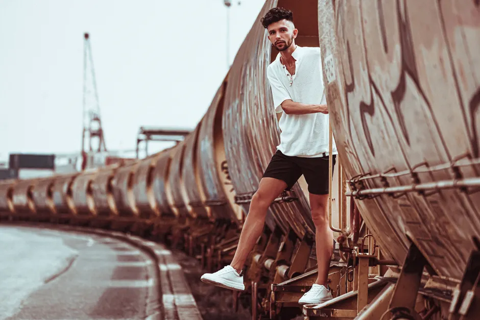 Homme en short noir et chemise blanche posant dans un décor urbain industriel