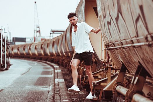 Stylish man in casual attire posing alongside industrial train cars.