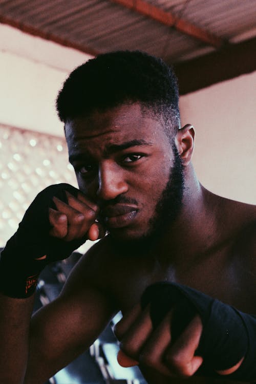 Powerful African American male with beard with hand near face looking at camera while practicing boxing techniques in gym in daylight