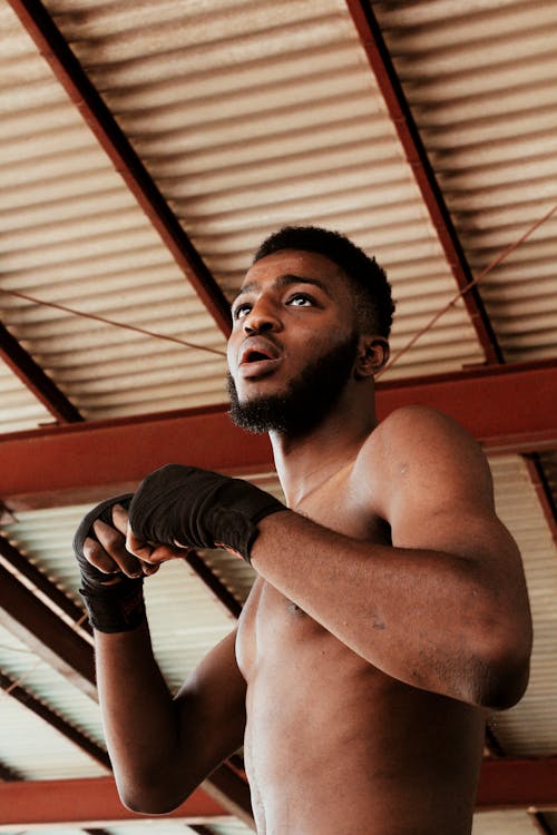 Low angle of African American male boxer with opened mouth in bandages looking away while training in spacious room in daytime