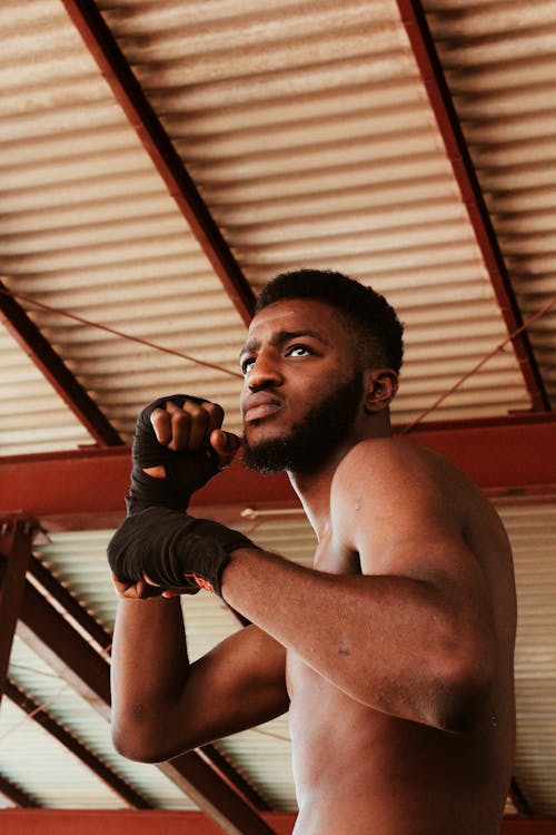 From below of aggressive bearded shirtless African American male fighter with hand near face practicing boxing punches while standing in stance