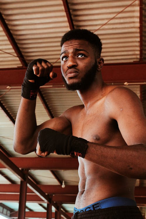 From below of African American male boxer with bandage on fists with naked torso looking away while training technique in gym