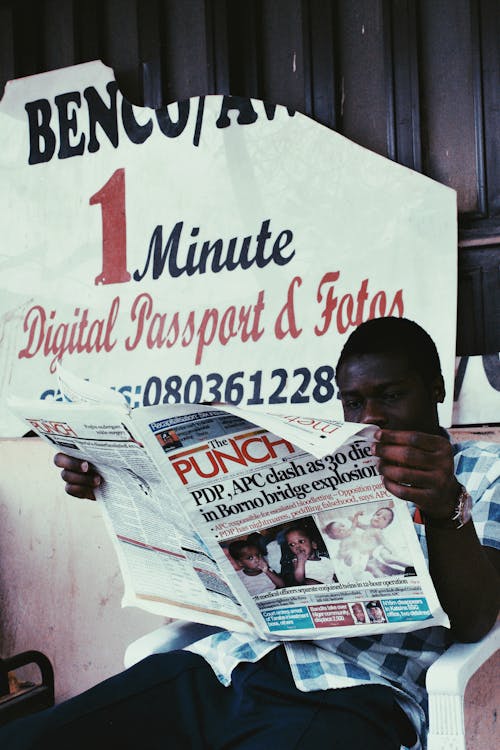 Focused anonymous African American male with newspaper covering half face sitting in chair near wall with torn poster in daylight