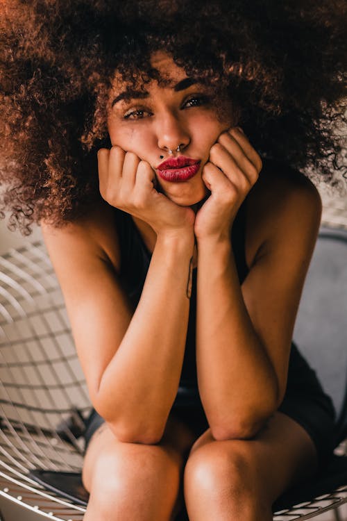 Crop melancholic ethnic female with Afro hair sitting on chair and holding face in hands while looking at camera with sadness