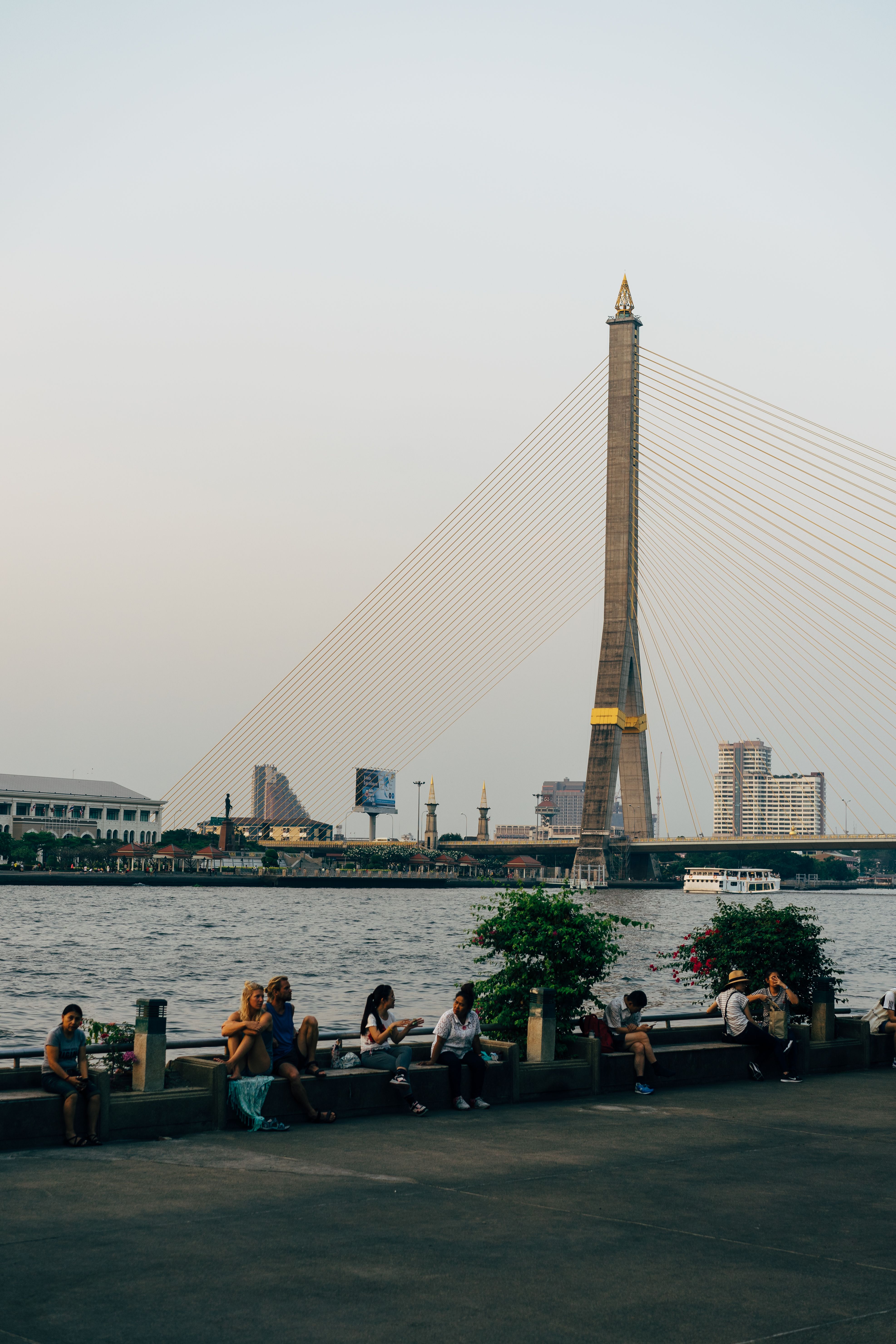 Outdoor scene of Rama VIII Bridge in Bangkok with cityscape and people along the riverfront.