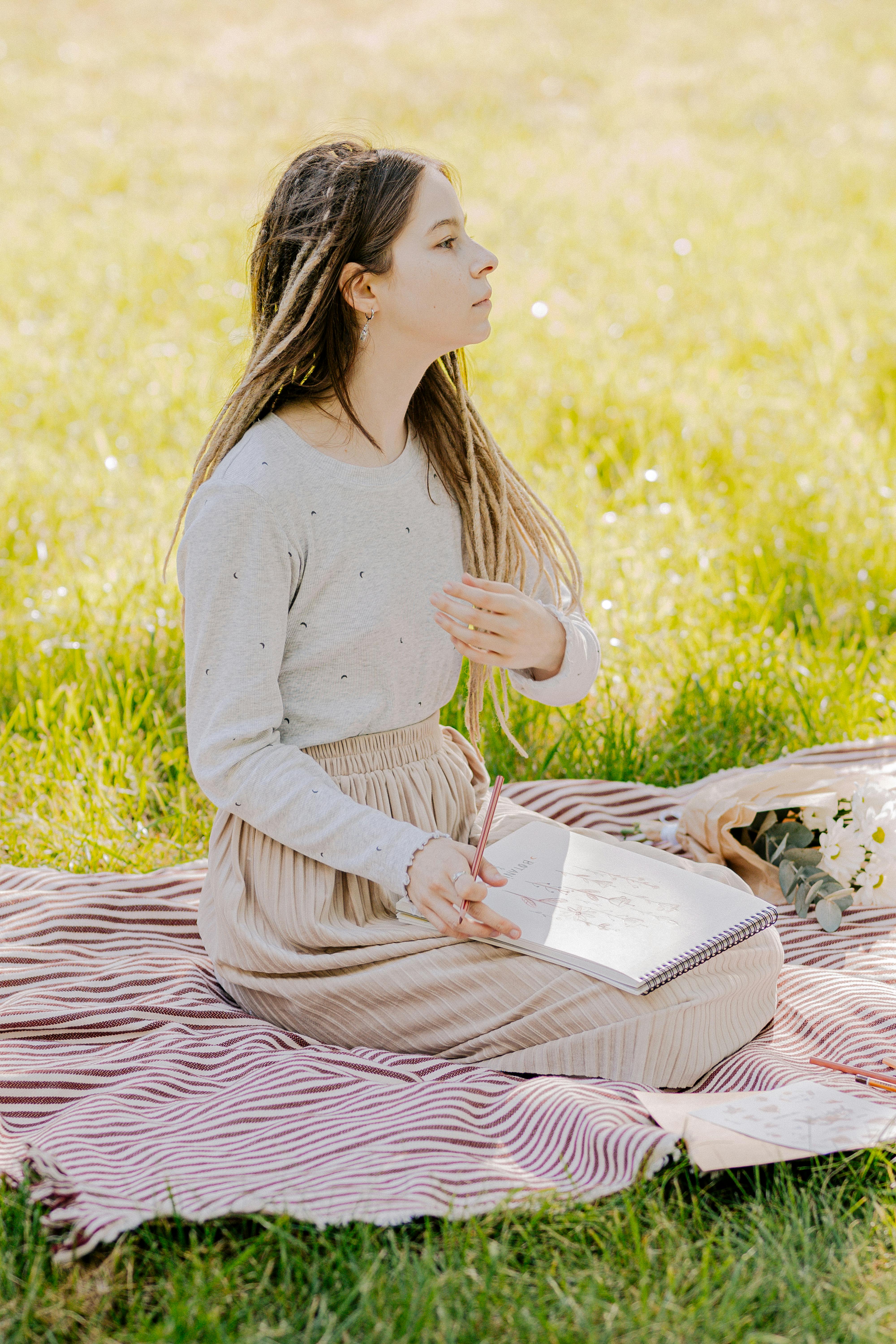 Delighted teen girl sitting in meadow and drawing in sketchbook