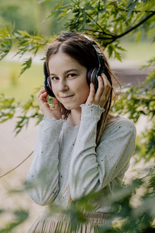 Free A young woman in a gray sweater listening to music with headphones outdoors, surrounded by greenery. Stock Photo