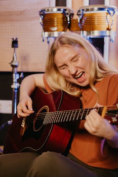Blonde woman intensely playing acoustic guitar with drums in background, showcasing musical passion.