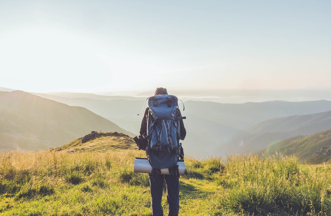 Back view anonymous male tourist with backpack standing on grassy hilltop and admiring magnificent mountainous landscape on sunny summer day